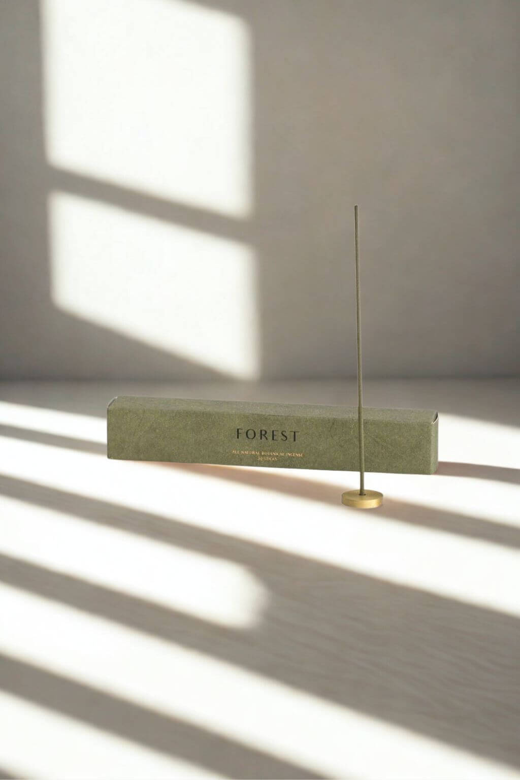 Minimalist display of a burning Hinoki incense stick in a brass holder, shadows of a window falling across the scene beside the green "Forest" box.