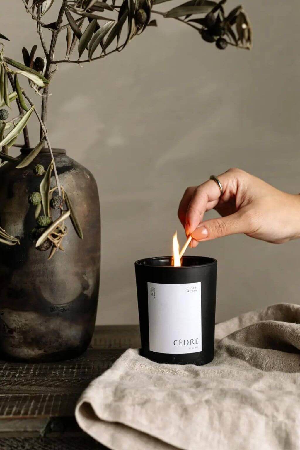 Hand lighting the Cedar Luxury Candle with a match, styled with an olive branch in a ceramic vase.