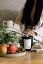 Cedar Luxury Candle burning on a wooden table, with apples, tomatoes, and evergreen sprigs nearby.