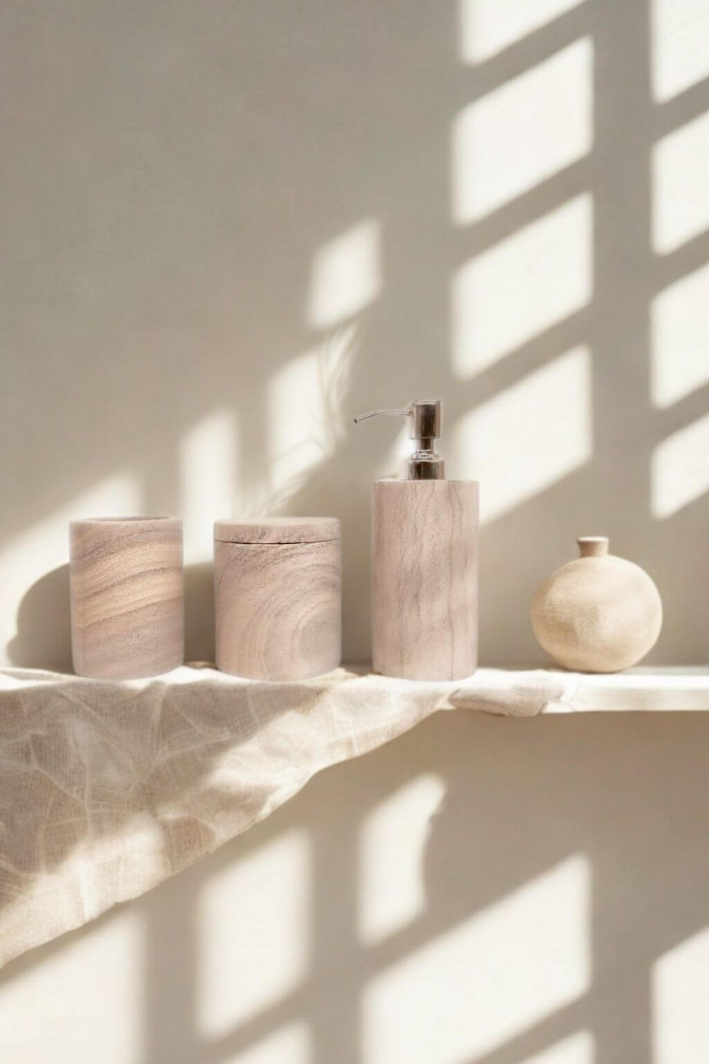 Rainbow sandstone soap dispenser, jar, and tumbler styled on a linen-draped shelf beside a ceramic vase, with soft shadows falling across the wall.