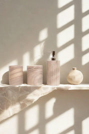 Rainbow sandstone soap dispenser, jar, and tumbler styled on a linen-draped shelf beside a ceramic vase, with soft shadows falling across the wall.