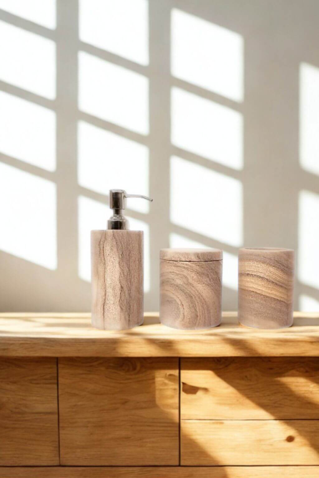A trio of rainbow sandstone bath accessories on a wooden counter, including a soap dispenser, covered jar, and tumbler, bathed in sunlight through a window.