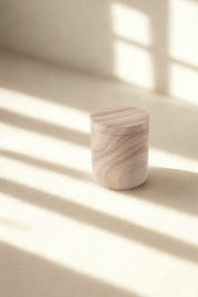 Round sandstone storage jar with fitted lid, crafted from rainbow sandstone, highlighted by soft windowpane shadows.