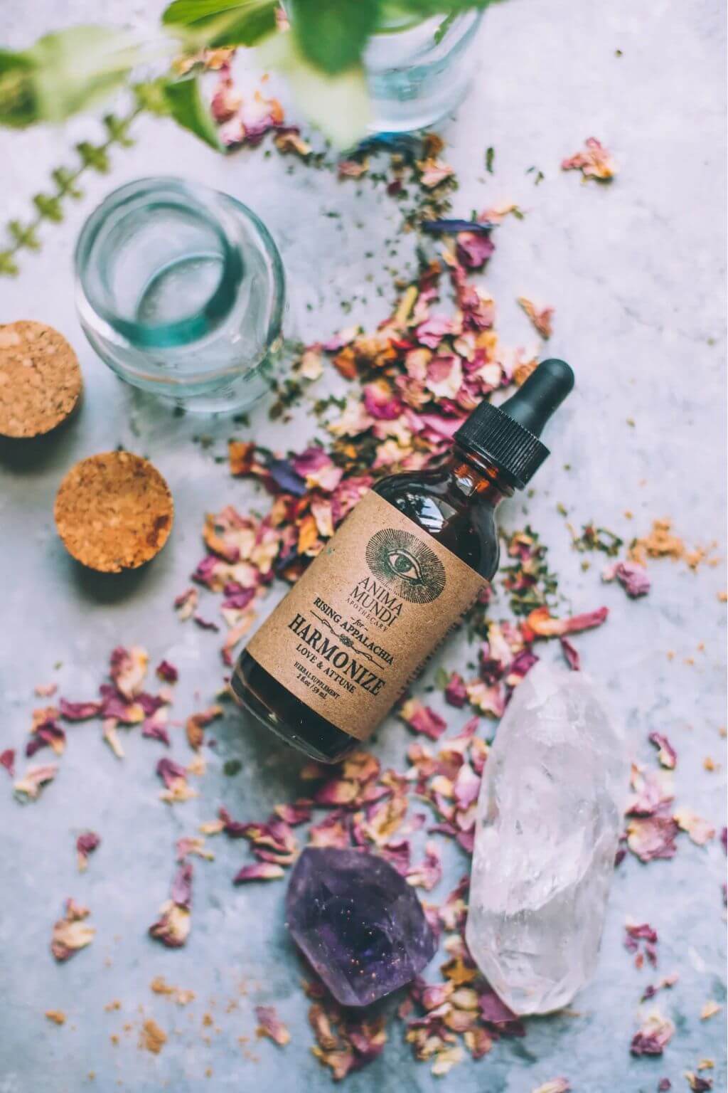 A bottle of Anima Mundi Harmonize Tonic on a gray background with rose petals and crystals in the background