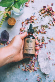 Hand holding a bottle of Anima Mundi Harmonize Tonic with rose petals and crystals in the background