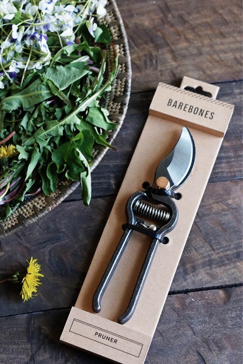 Pruner in Barebones packaging on a wooden surface with a bowl of greens and flowers.
