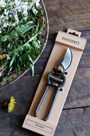 Pruner in Barebones packaging on a wooden surface with a bowl of greens and flowers.