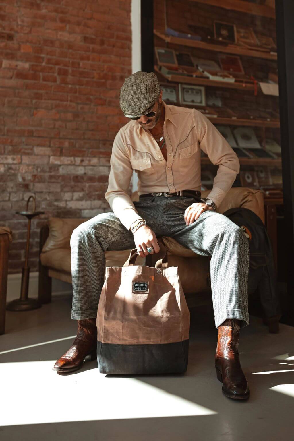 Man sitting on a chair holding a brown leather bag in a room with brick walls and wooden furniture.