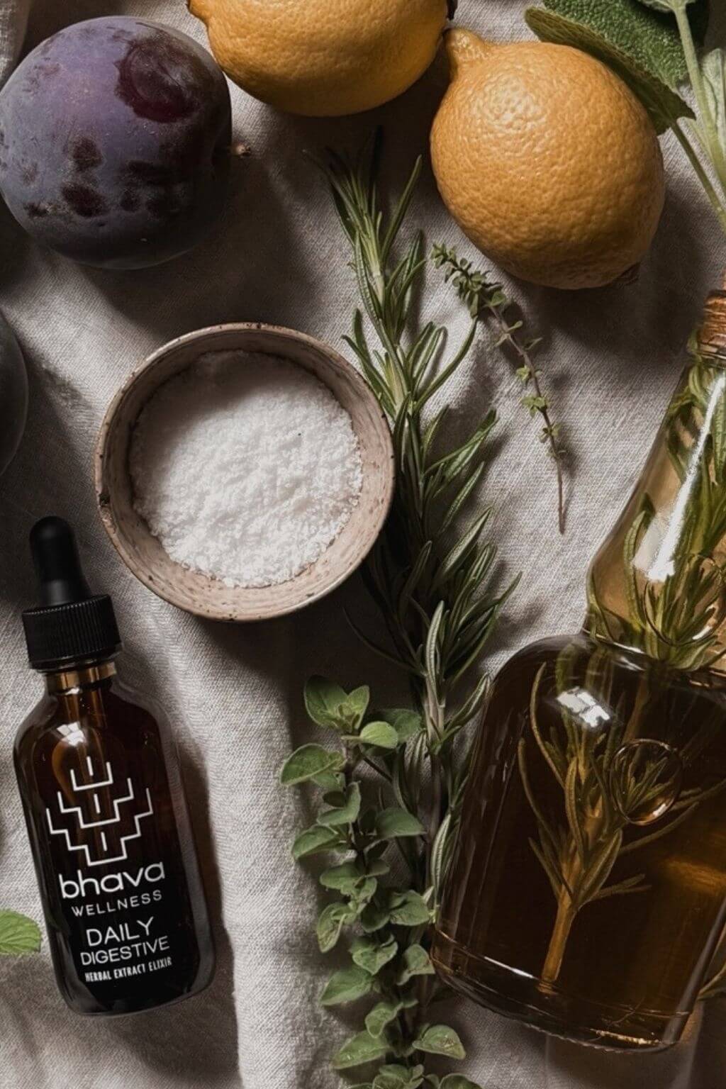 Flat lay imagee of Bhava Wellness Daily Digestive Herbal Tincture on a linen cloth with plums, lemons, fresh herbs, a glass bottle of rosemry infused oil nd a ceramic bowl of sea salt