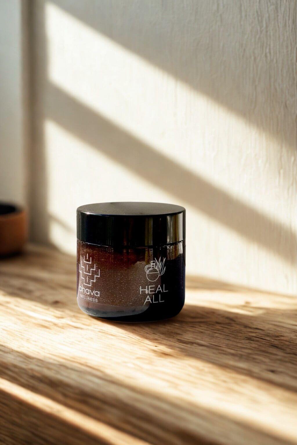 Amber glass jar of Heal All Herbal Salve by Bhava Wellness on a wooden surface with sunlight casting shadows.