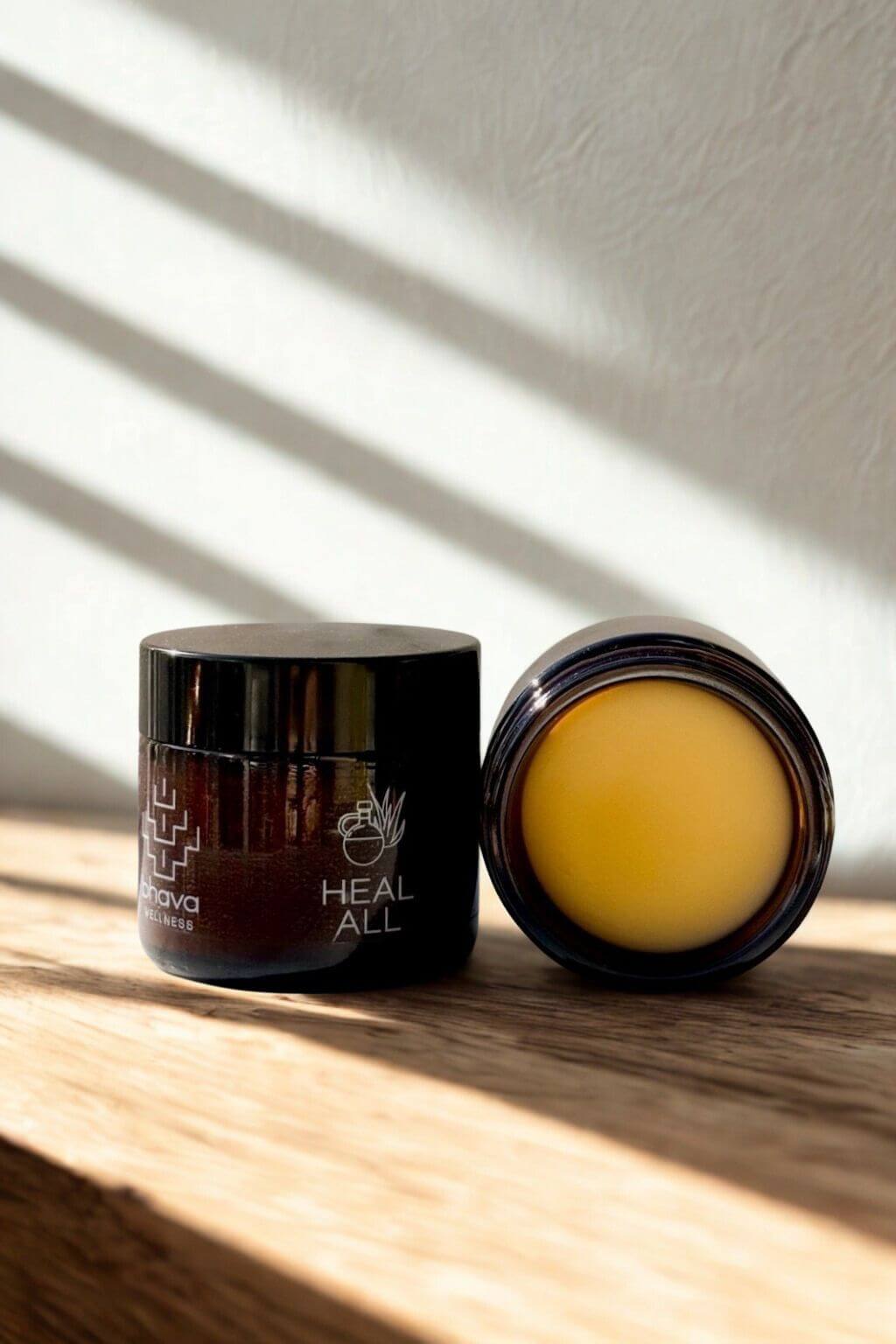 Two jars of Heal-All Herbal Salve, one open showing a vibrant yellow balm, on a wooden surface with sunlight casting shadows.