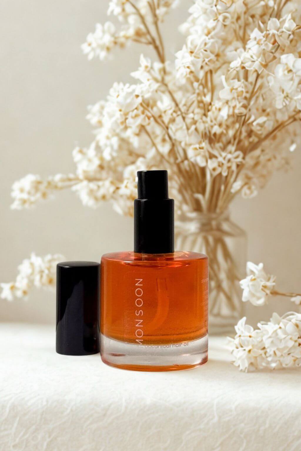 Elegant bottle of Monsoon Botanical Hair Oil with cap off alongside delicate white flowers in a glass vase