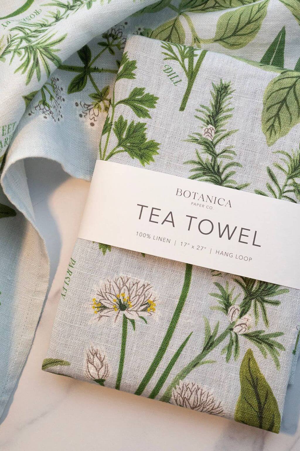 Herb Garden Linen Tea Towel in packaging displayed on a marble surface, surrounded by the same linen fabric printed with green botanical illustrations.