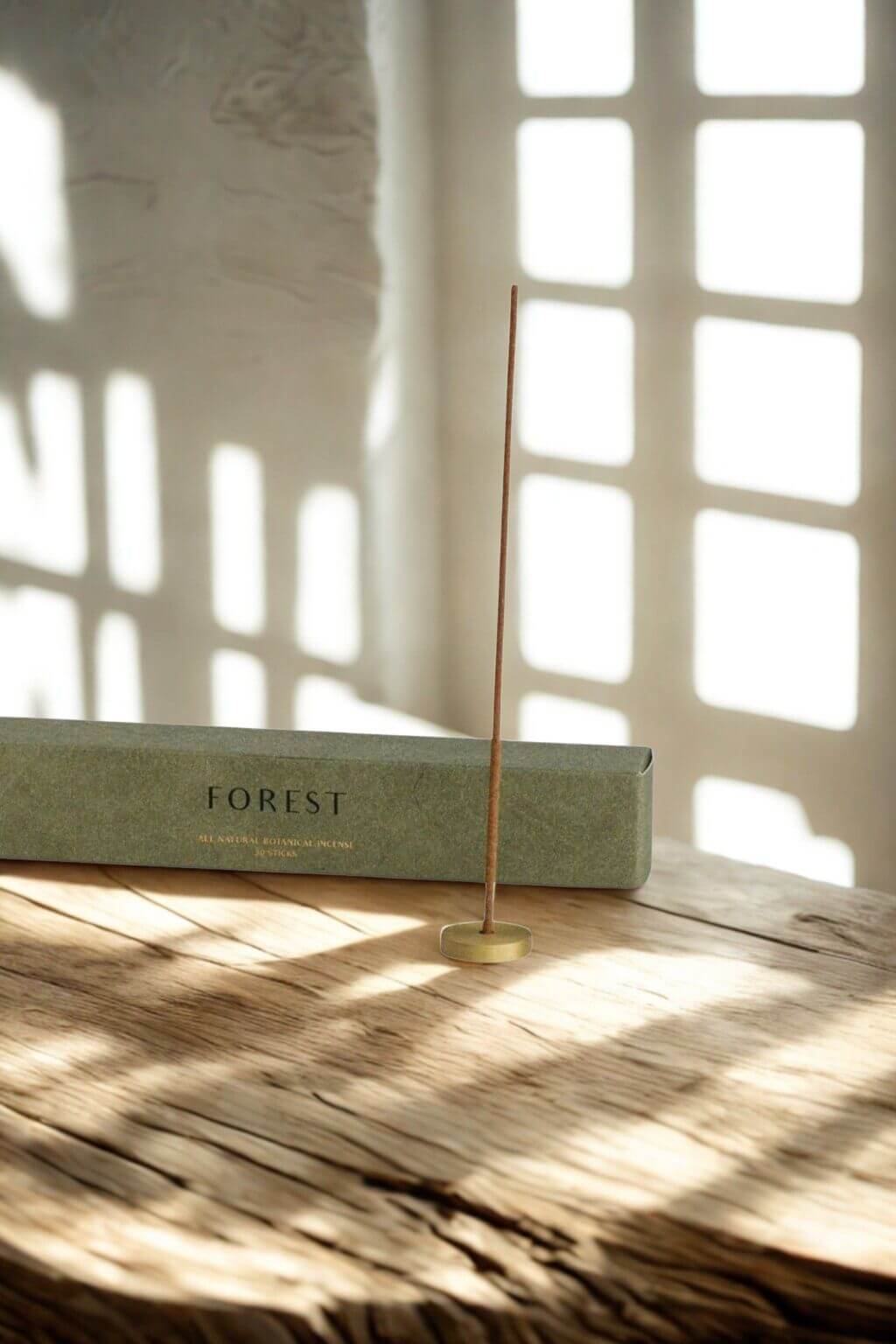 A single cedarwood incense stick burning in a brass holder on a rustic wooden surface, soft sunlight streaming through a window in the background.
