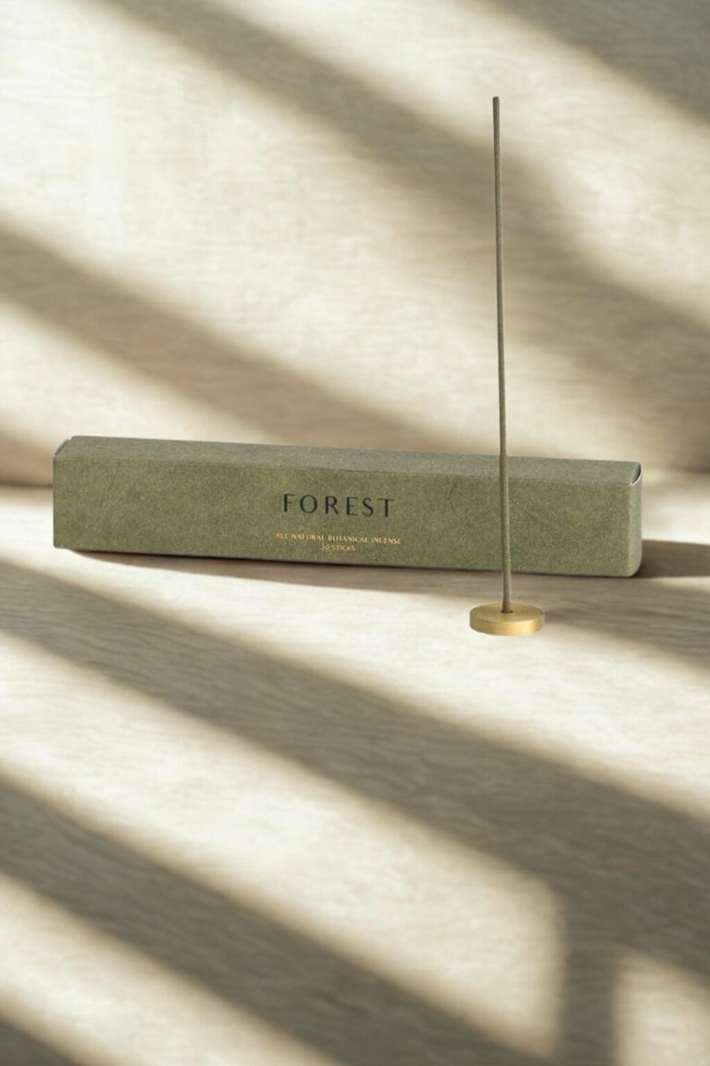 A single Hinoki Japanese incense stick standing upright in a brass holder, with the green "Forest" box in the background, bathed in soft natural light.
