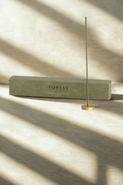 A single Hinoki Japanese incense stick standing upright in a brass holder, with the green "Forest" box in the background, bathed in soft natural light.