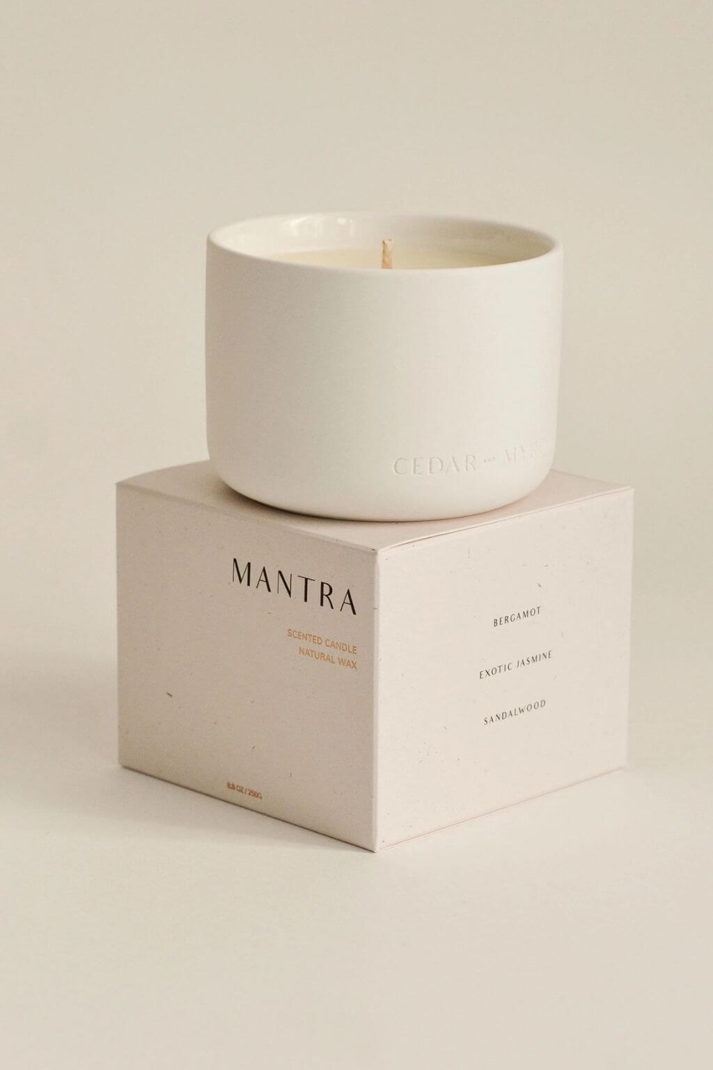 A Cedar & Myrrh Mantra Wellness Ritual Candle in a matte white ceramic vessel, resting atop its minimalist cream box, with fragrance notes listed on the side.