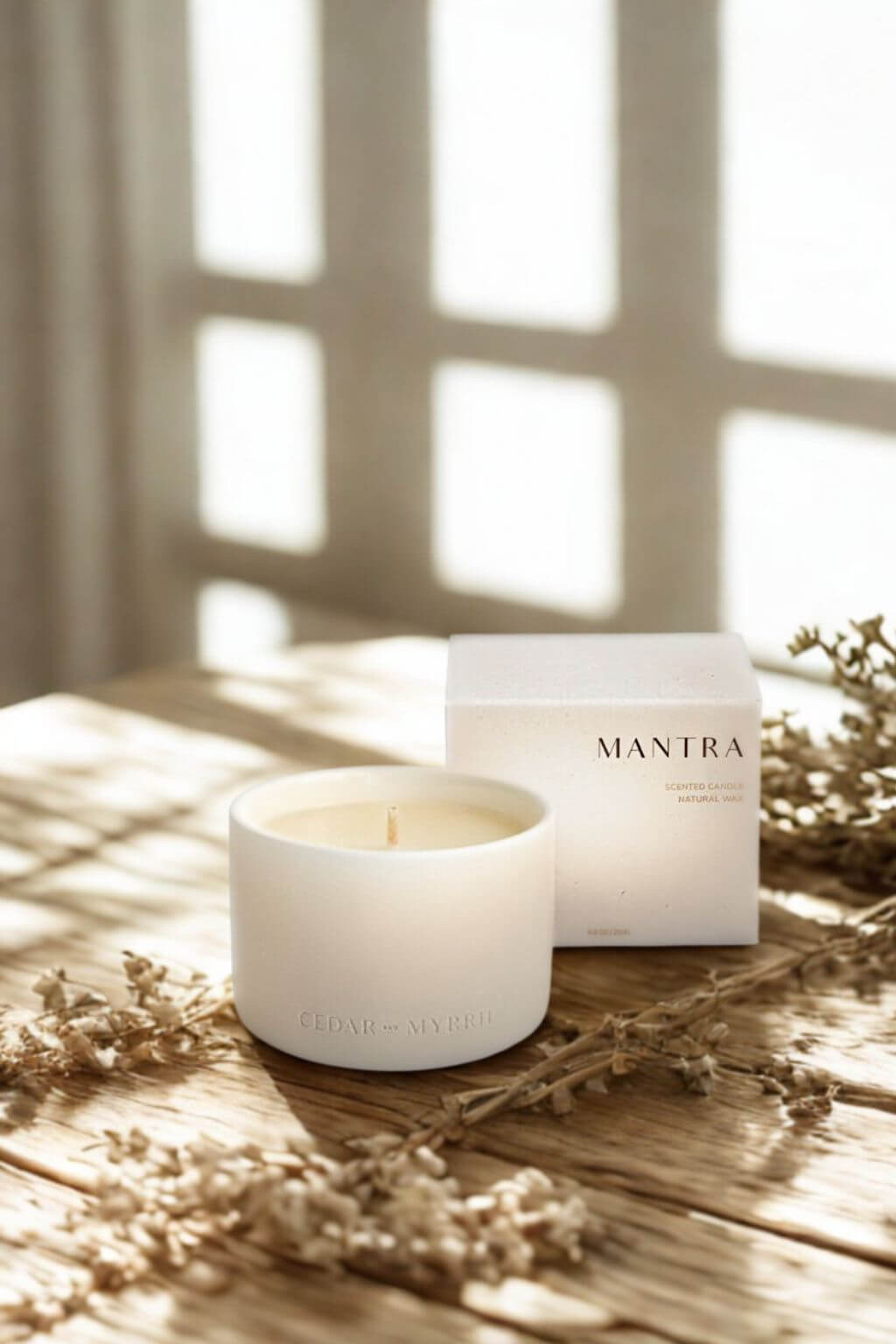 Mantra Wellness Ritual Candle displayed on a rustic wooden surface, surrounded by delicate dried stems, with sunlight filtering through windowpanes.
