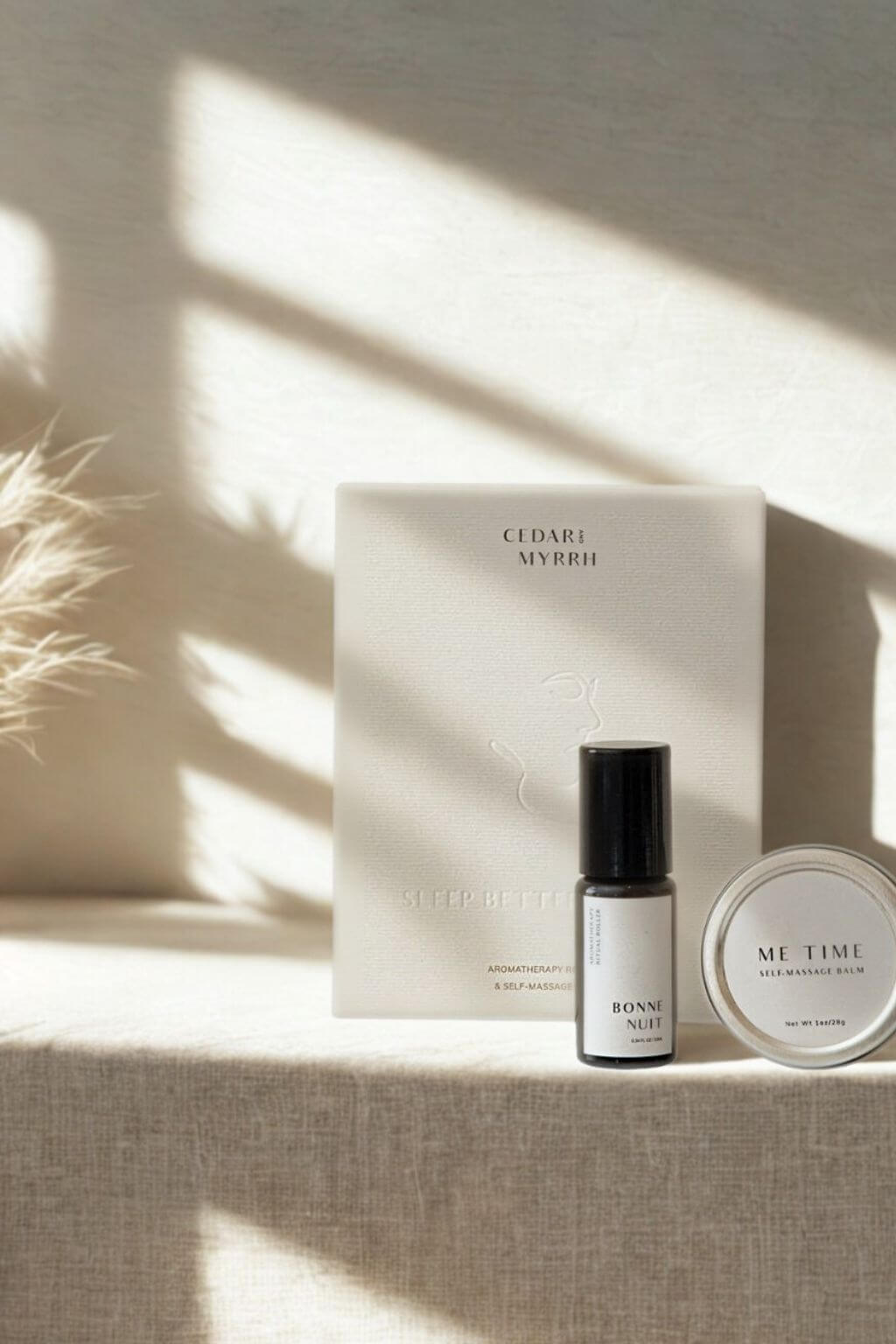 Cedar + Myrrh roller bottle of Bonne Nuit, and Me Time Balm with embossed gift box on a textured surface with sunlight casting shadows