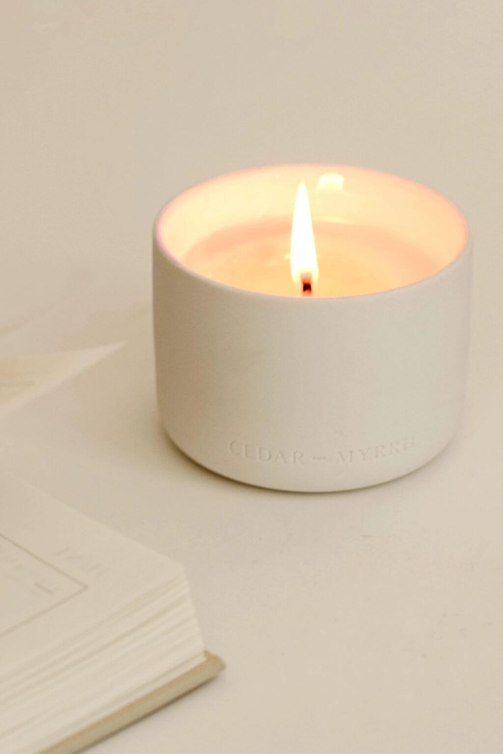 Close-up of a burning Deep Breath candle in a white ceramic vessel, placed on a white tabletop with an open book.