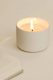 Close-up of a burning Deep Breath candle in a white ceramic vessel, placed on a white tabletop with an open book.