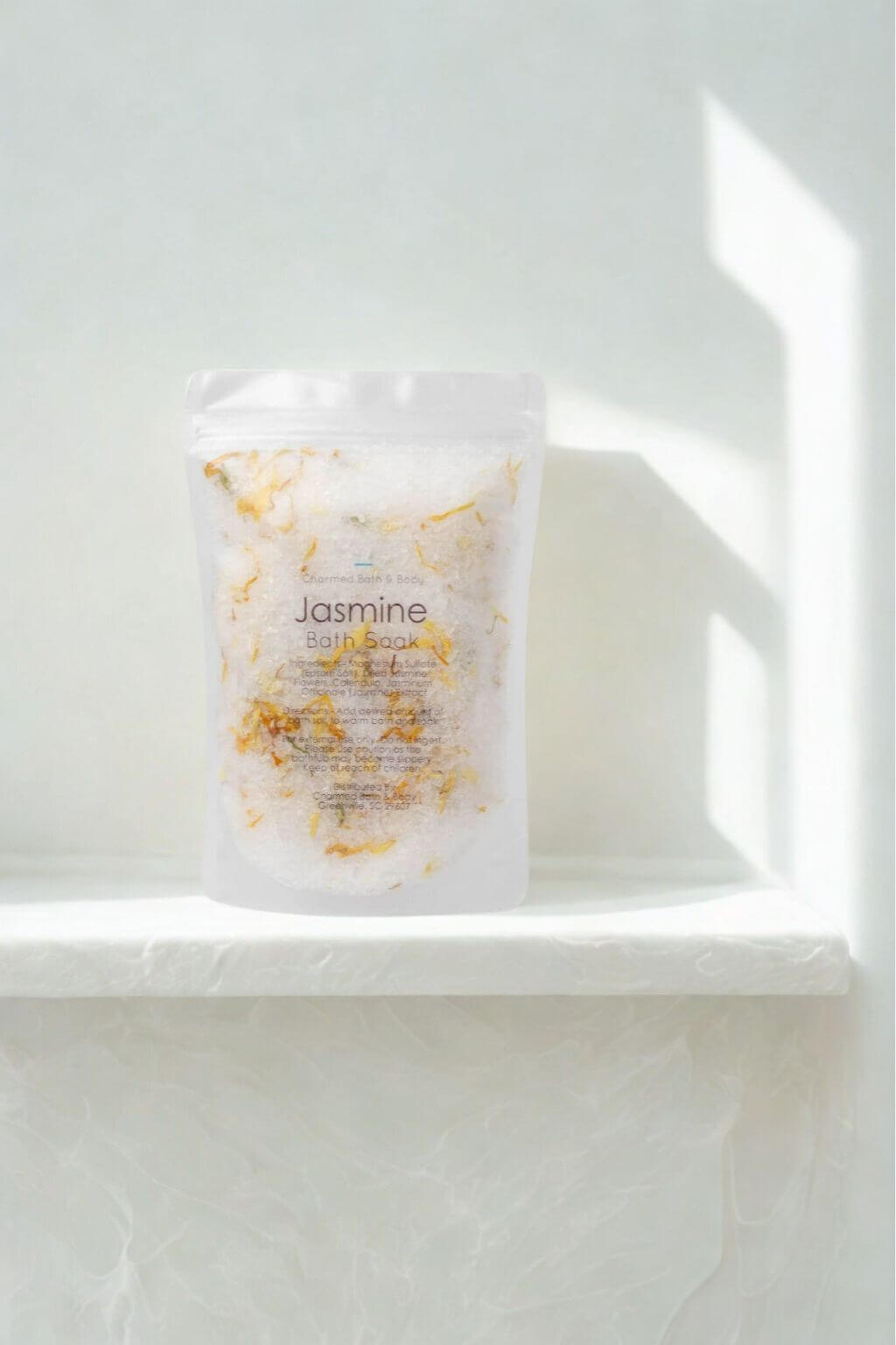 Clear matte pouch of Charmed Handmade Jasmine Bath Soak displayed on a white ledge with soft natural light casting shadows across the background.