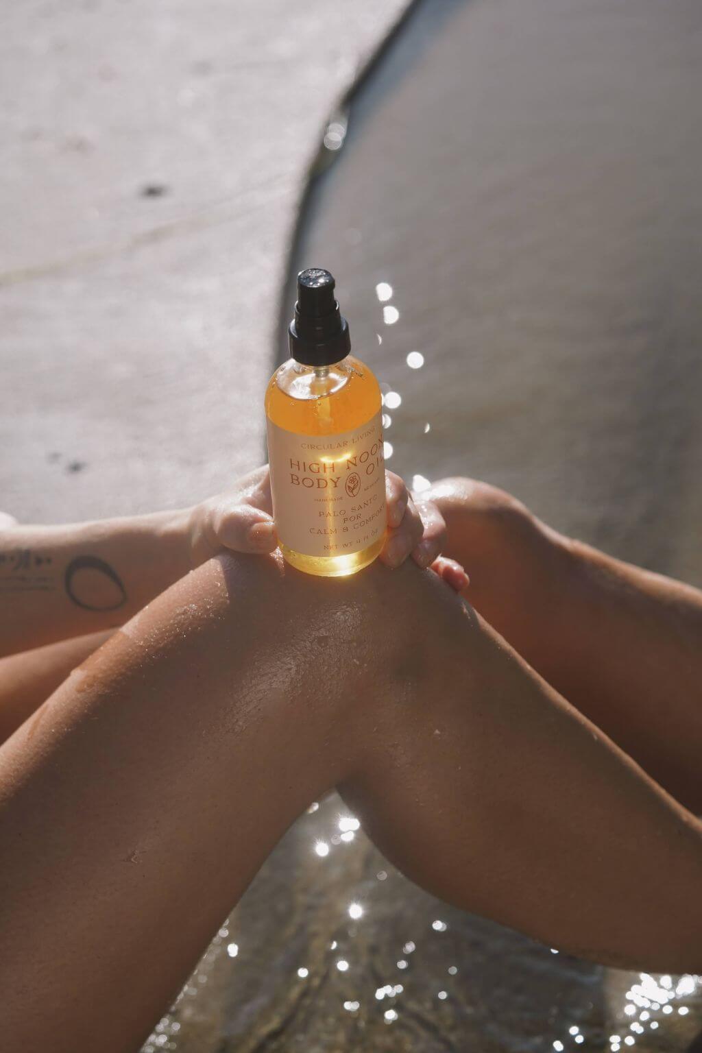 High Noon Body Oil bottle held on a woman's knee, resting by water, reflecting sunlight