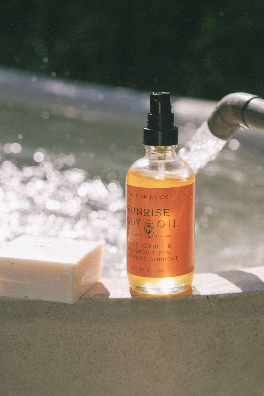 Sunrise Body Oil resting on ledge beside natural soap near flowing water.