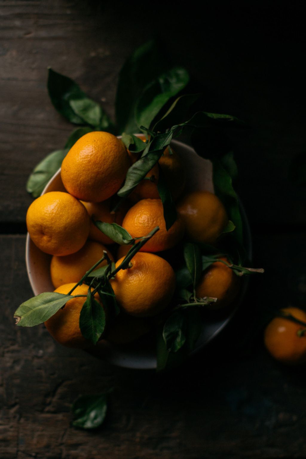 A rich, moody image of a bowl of fresh citrus with dark green leaves set against a dark walnut wood table