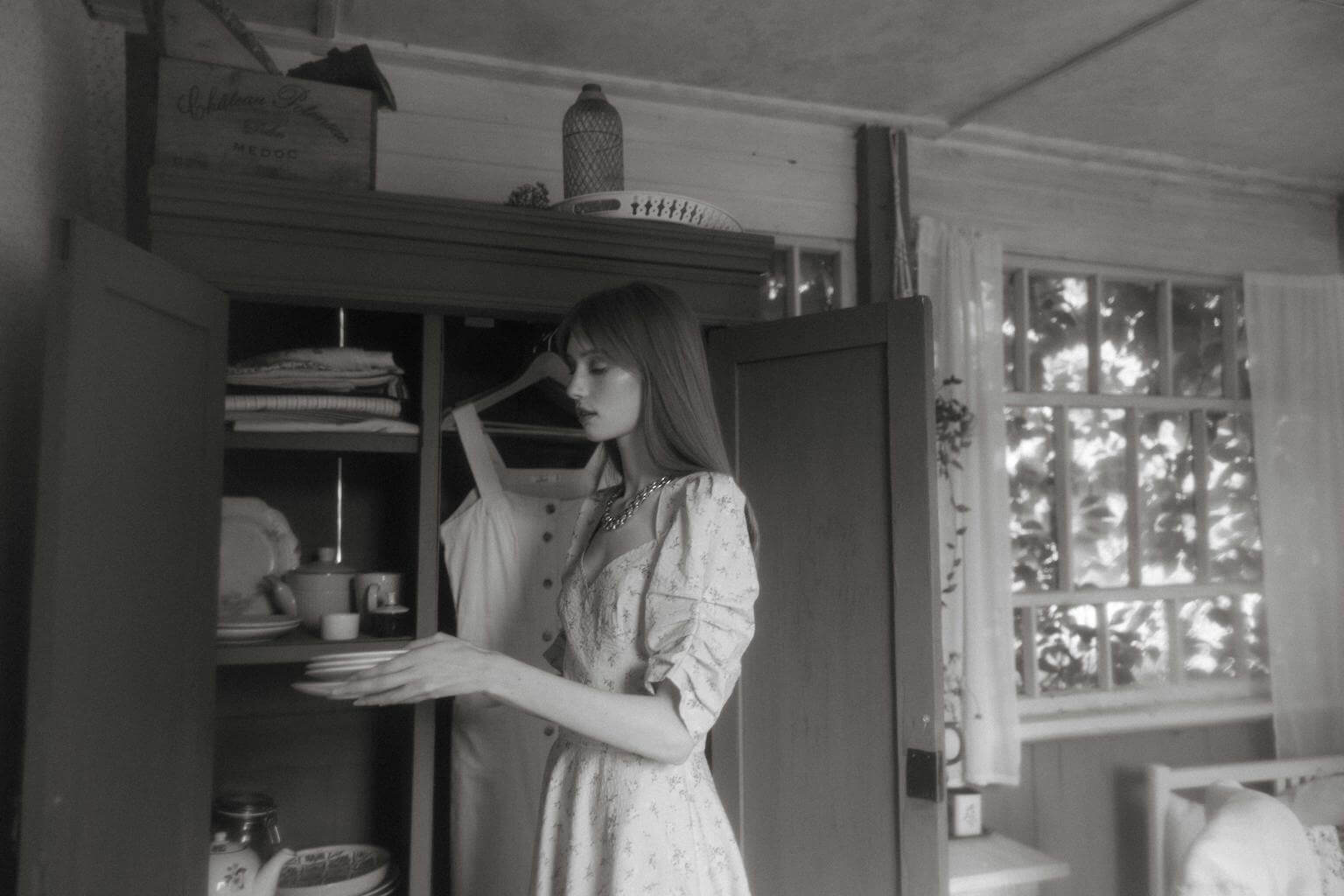 BW image of a woman in a floral peasant dress looking through an antique hutch and admiring her vintage wares