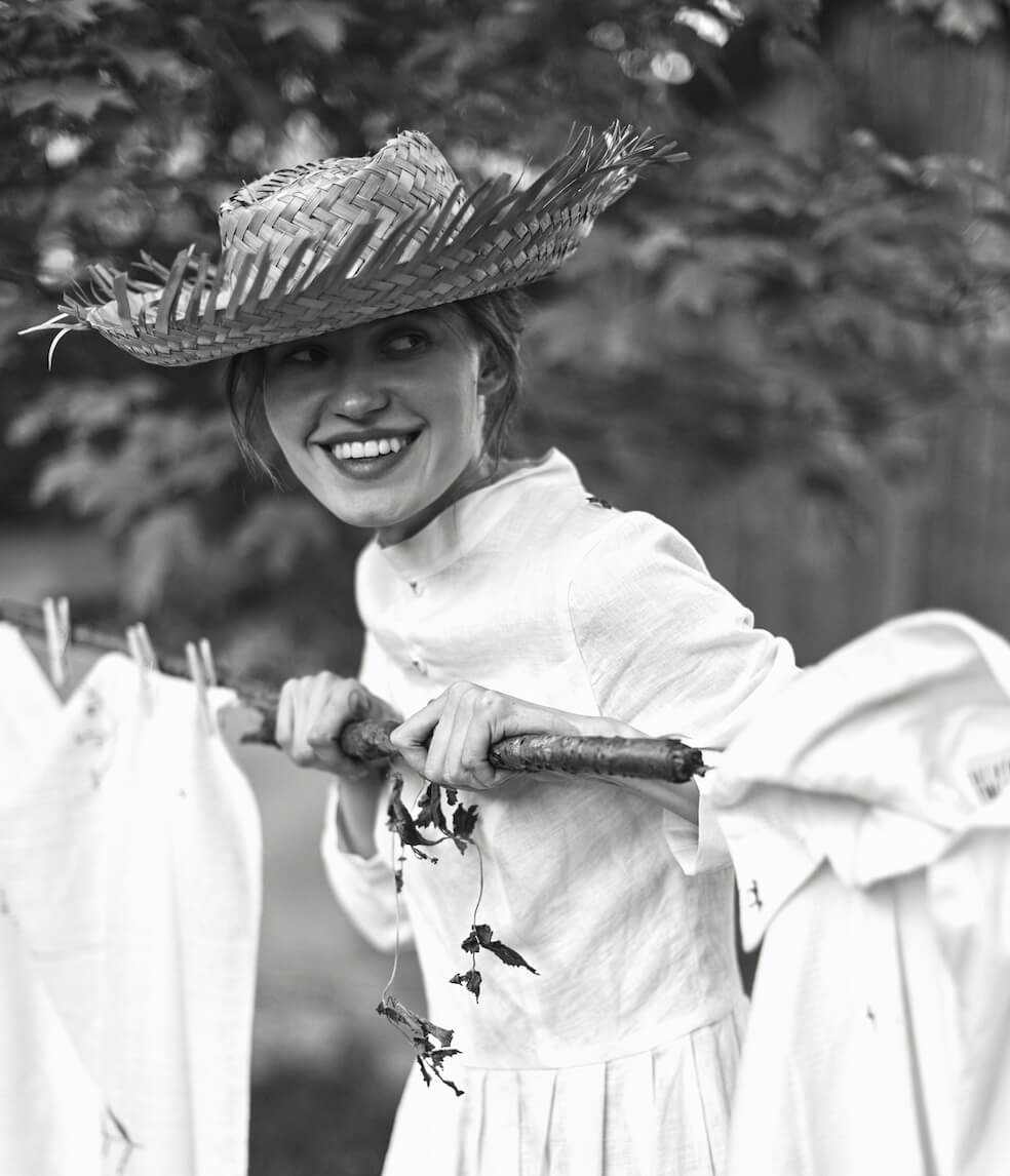 young woman with a straw hat and a  long white dress standing beneath a tree in front of a clothesline hung with white linens