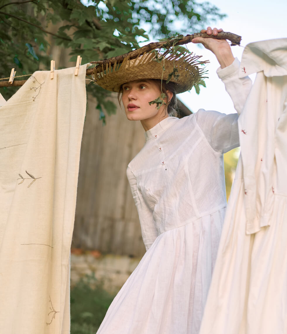 woman with white dress and straw hat standing behind a clothing line strung with neutral linens