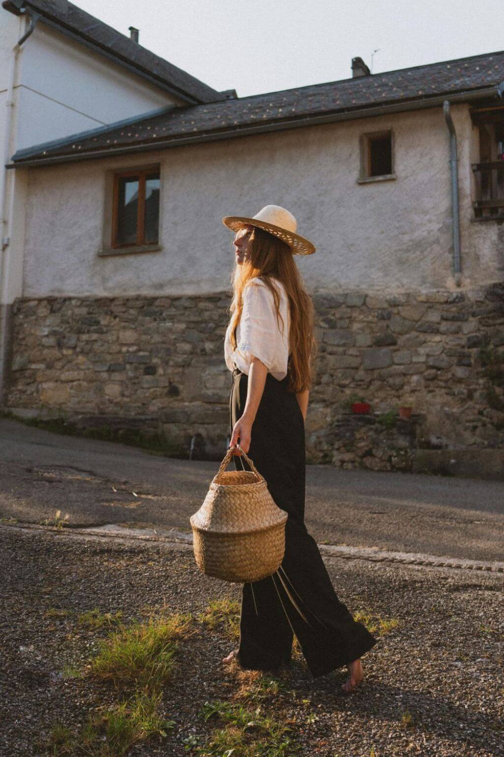 young woman wearing a straw hat, white poet's blouse and corduroy trousers hollding a market basket in a small european villlage