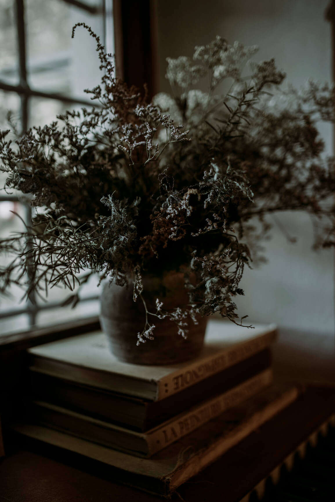 dark moody photo of dried flowers and evergreens atop a stack of books with a mullioned window in the background