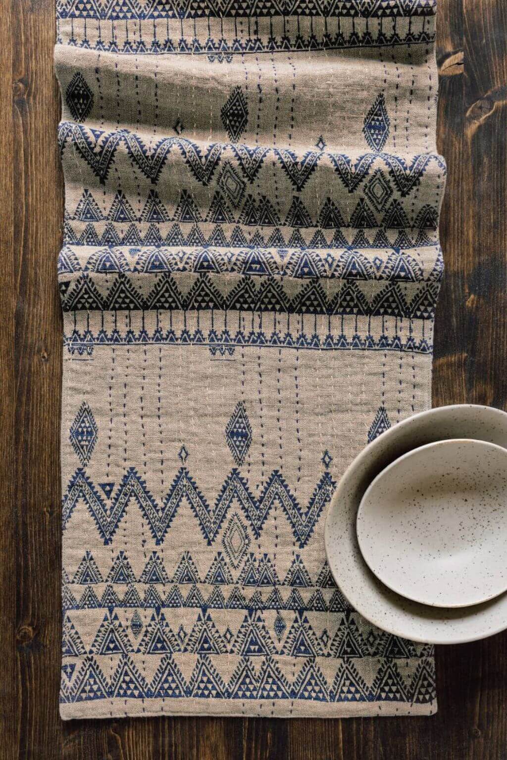 Enigma table runner in tan linen with hand-stitched blue Kantha embroidery, styled across a rustic wooden table.