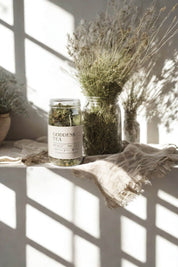 Apothecary setting with Evergreen Apothecary Goddess Tea jar beside dried herbs and woven linen