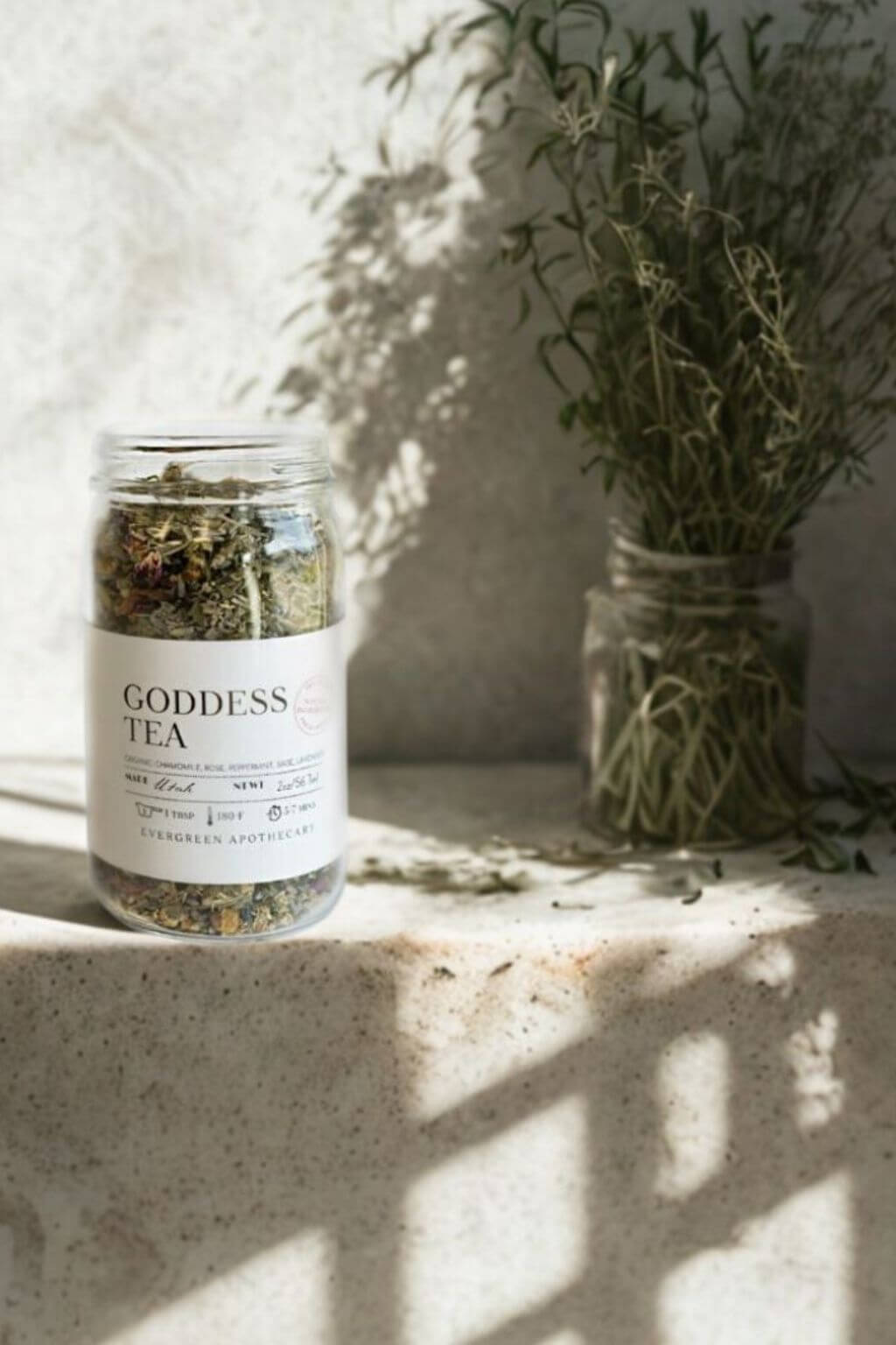 Glass jar of Evergreen Apothecary Goddess Tea on stone ledge with herbs in the background