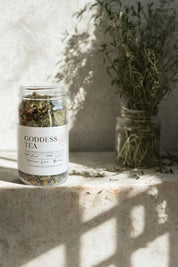 Glass jar of Evergreen Apothecary Goddess Tea on stone ledge with herbs in the background