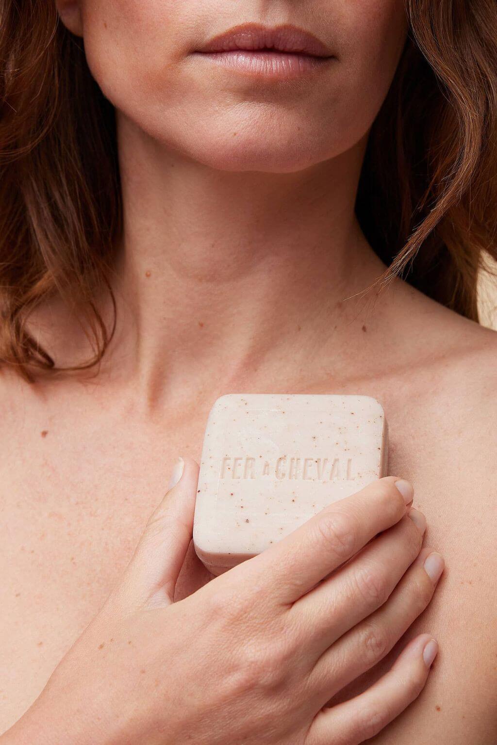 Woman holding Fer à Cheval Gentle Cleansing Soap bar against her collarbone.