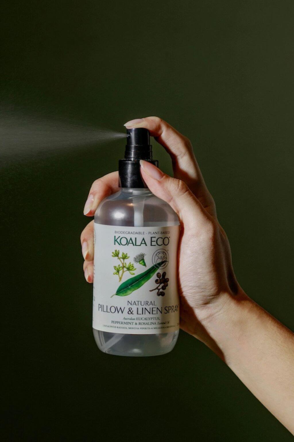 Hand holding a bottle of Koala Eco's Natural Pillow and Linen Spray against a dark green background