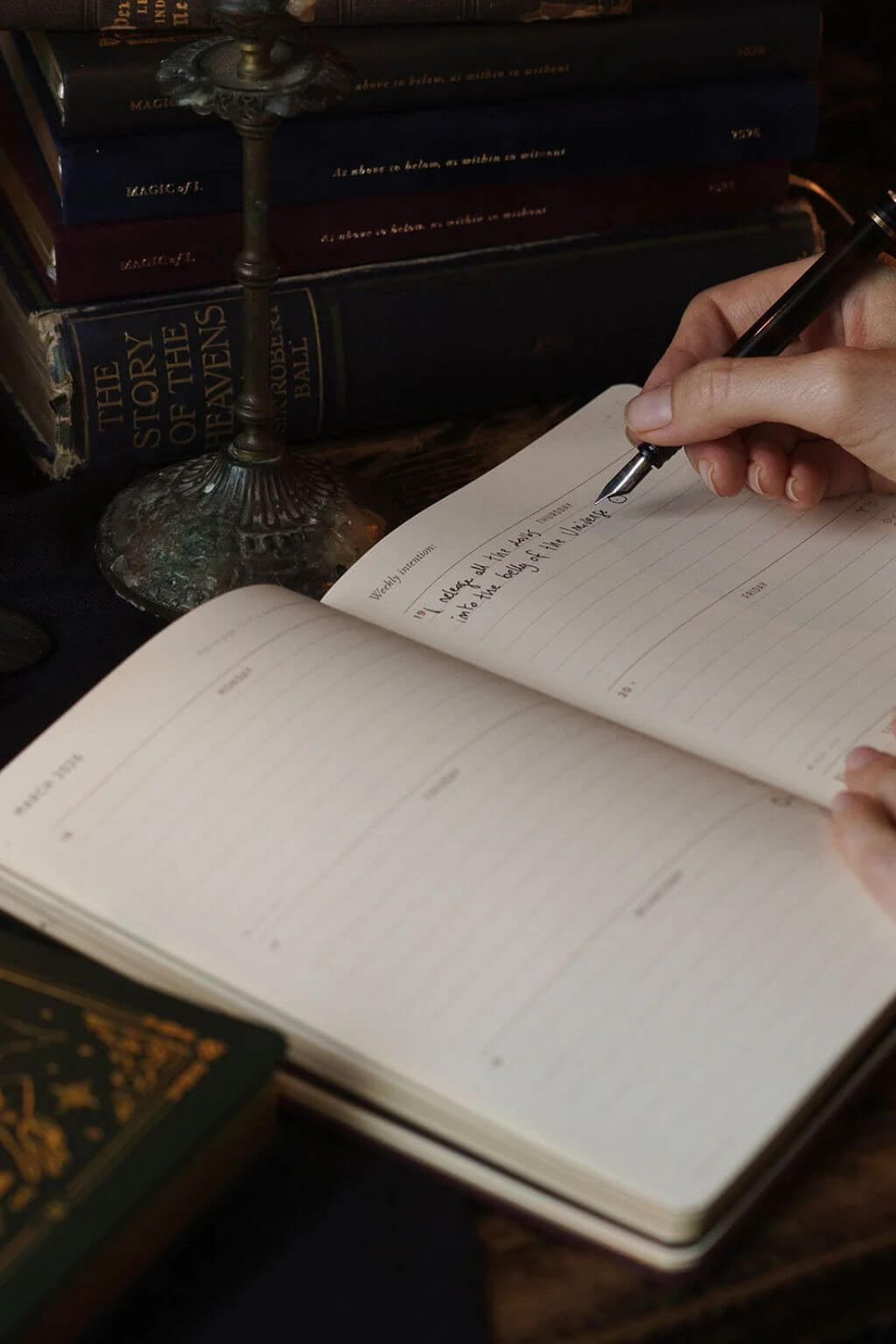 Hand writing in a Magic of I 2026 planner iwith books stacked in the background