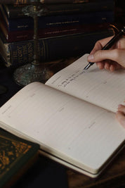 Hand writing in a Magic of I 2026 planner iwith books stacked in the background