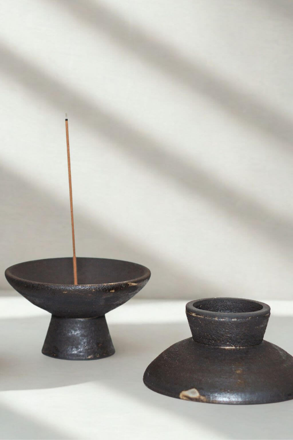 Raw black stoneware incense holders in shibui style, one holding a lit incense stick, bathed in soft natural light.
