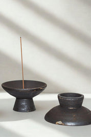 Raw black stoneware incense holders in shibui style, one holding a lit incense stick, bathed in soft natural light.