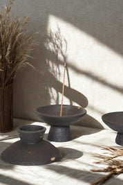 Black stoneware incense holders arranged beside dried botanicals, shadows falling across a linen background.