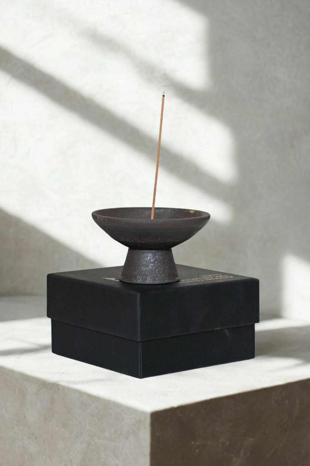 Shibui stoneware incense holder resting atop a matte black gift box, incense stick standing gracefully in morning light.