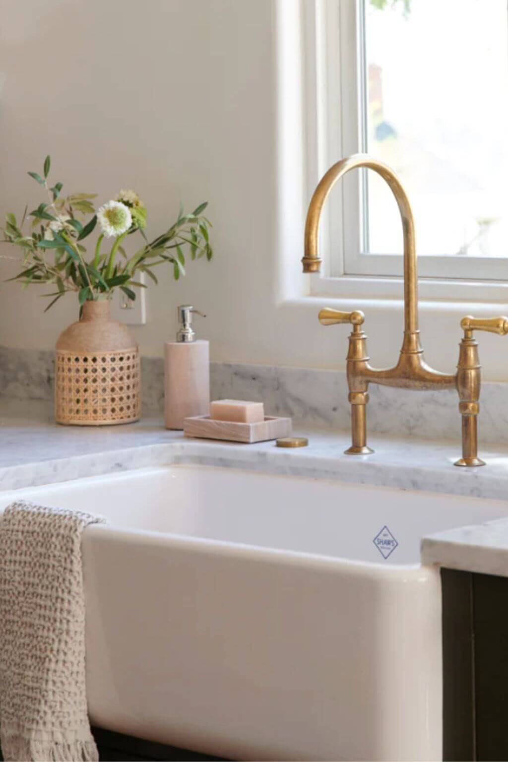 armhouse kitchen sink vignette with brass faucet, marble countertop, and the Rainbow Sandstone Soap Dispenser alongside a natural rattan vase and bar soap.