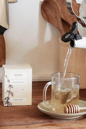 Water being poured into a glass mug with Juniper Ridge White Sage + Wild Mint Tea on a wooden surface